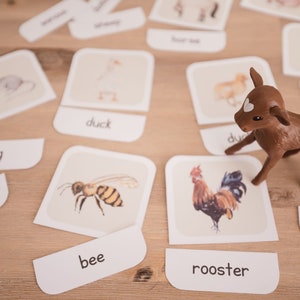 May include: A collection of animal flashcards and a brown toy deer are arranged on a light wooden surface. The flashcards feature illustrations of animals like a bee, duck, and rooster, with their names printed below. The toy deer stands near the cards.