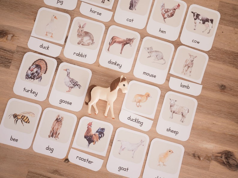 May include: Educational flashcards featuring farm animals, including a duck, rabbit, donkey, and more. A toy horse is placed among the cards. The cards have the animal's name printed below the image. The background is a light brown wooden surface.