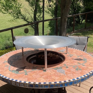 May include: A round, tiled patio table with a metal fire pit insert and a metal mesh cover. The table has a blue and white border and a brown and white tiled top.