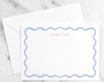 Op de afbeelding: Een witte kaart met een lichtblauwe golvende rand. De tekst "EMMA RAE" is in roze in het midden van de kaart gedrukt.