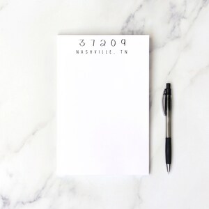 May include: A white notepad with the text "37209 NASHVILLE, TN" printed on the top. A black pen is lying on a white marble surface next to the notepad.