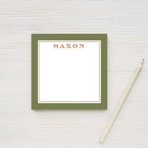May include: A green notepad with a white interior and a thin brown border. The word "SAXON" is printed in orange at the top of the notepad. A pencil is lying on a white wooden surface next to the notepad.