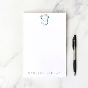 May include: A white notepad with a blue and floral monogrammed design. The notepad has the name "CANDICE JORDAN" printed at the bottom. A black pen is lying next to the notepad.