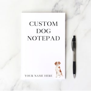 May include: A white notepad with the text "CUSTOM DOG NOTEPAD" and "YOUR NAME HERE" in black. A watercolor illustration of a dog is in the lower right corner. A black and silver pen is on the right side.
