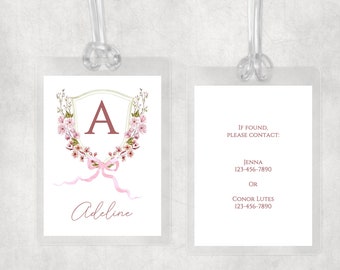 May include: A luggage tag with a pink floral wreath and a shield with the letter A in a burgundy color. The tag also has the name Adeline in burgundy script. The back of the tag has the text "If found, please contact:" followed by two names and phone numbers.
