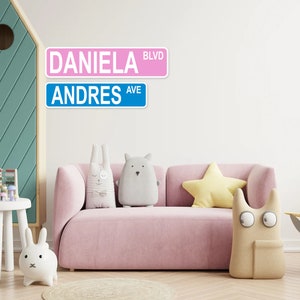 Puede incluir: Un letrero de calle rosa y azul con los nombres "Daniela Blvd" y "Andres Ave" sobre un fondo blanco. El letrero est&aacute; montado en una pared blanca sobre un sof&aacute; rosa con una almohada de estrella amarilla y un animal de peluche gris y blanco.