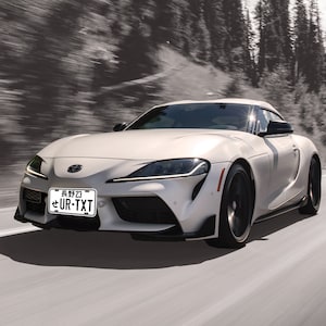 May include: A white Toyota Supra sports car with black wheels and a black number plate that reads "UR-TXT". The car is driving on a road with trees in the background.