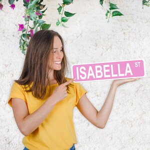 Puede incluir: Una mujer se&ntilde;ala una se&ntilde;al de calle rosa con el nombre "ISABELLA ST" escrito en letras blancas.