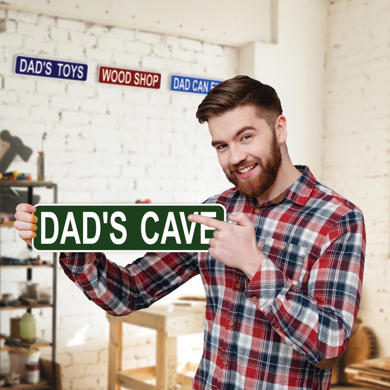 Puede incluir: Un hombre con una camisa a cuadros rojos y negros sostiene un letrero verde que dice "DAD'S CAVE". El letrero es de estilo se&ntilde;al de tr&aacute;fico con letras blancas. Hay otros letreros en el fondo que dicen "DAD'S TOYS", "WOOD SHOP" y "DAD CAN FIX IT".