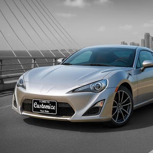 May include: Silver sports car with black rims and a front license plate that reads "3 Lines to Customize Your Ride". The car is parked on a paved road with a bridge and city skyline in the background.