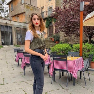 Puede incluir: Una mujer con una camiseta blanca, una blusa de sat&eacute;n verde, pantalones vaqueros negros y botines negros con tachuelas. Est&aacute; de pie en un patio con mesas y sillas.