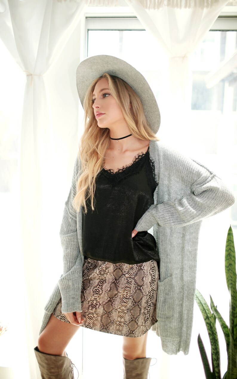 Puede incluir: Una mujer con un c&aacute;rdigan gris, una blusa negra de encaje, una falda de estampado de serpiente, un fedora gris y botas marrones.