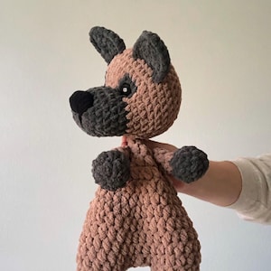 May include: A handmade crochet plush toy shaped like a dog, featuring a light brown body with dark gray ears, muzzle, paws, and feet. The toy has a black stitched nose and small dark eyes, crafted from a soft, chunky yarn with a visible looped texture.