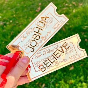 Puede incluir: Dos boletos dorados de Polar Express Round Trip con los nombres "Joshua" y "Believe" en letras brillantes. Cada boleto tiene un número único y está atado con una cinta roja sobre un fondo de hierba verde.
