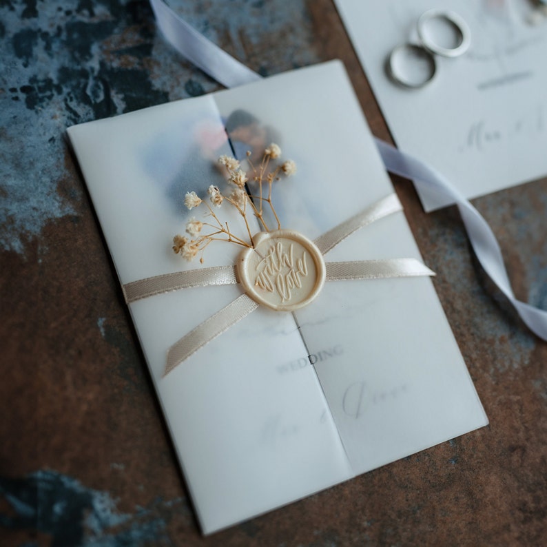 Pu&ograve; includere: Un invito di nozze bianco con un sigillo di cera che dice "with love" e un rametto di fiori secchi. L'invito &egrave; legato con un nastro beige.