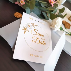 Pu&ograve; includere: Carta bianca salva la data con lettere dorate. La carta dice "Melisa & Michael Save the Date 02.18.22" con un cerchio dorato e un design floreale.