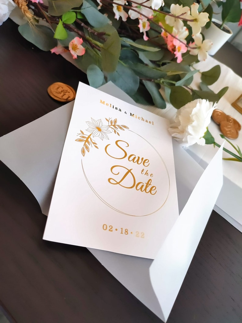 Pu&ograve; includere: Carta bianca salva la data con lettere dorate. La carta dice "Melisa & Michael Save the Date 02.18.22" con un cerchio dorato e un design floreale.