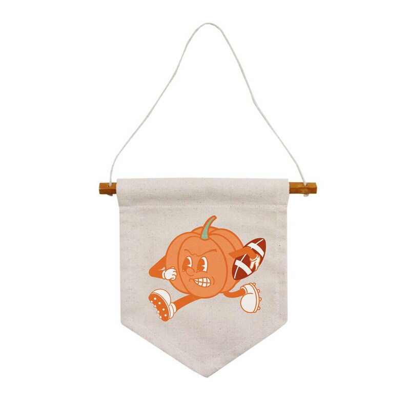 May include: A white canvas banner with a cartoon pumpkin wearing a football helmet and holding a football. The banner is hanging from a wooden dowel rod with a white string.