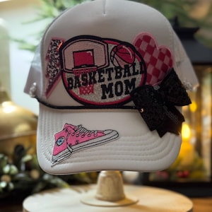 May include: White trucker hat with a mesh back, featuring a basketball-themed design. The front of the hat displays a pink basketball hoop, a basketball, and the text "BASKETBALL MOM." Additional embellishments include a pink shoe patch, a lightning bolt, a heart, and a black sequin bow.