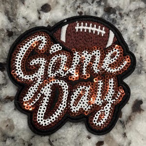 以下が含まれることがあります： 白と銅色のスパンコールでGame Dayの文字が施された装飾用ワッペン。文字の上には、白いレースが付いた刺繍の茶色のフットボールがデザインされています。全体は布地の上に太い黒い縁取りで囲まれています。