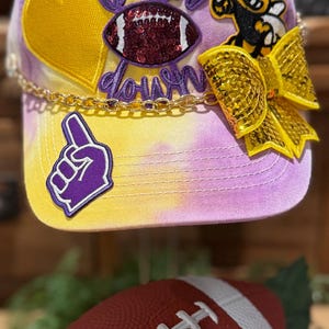 Könnte beinhalten: Eine Baseballkappe im Batik-Design in Gelb, Lila und Weiß mit den Worten "Touch Down" in lila Schrift. Die Kappe hat ein goldenes Herz, einen Football, eine Biene und eine goldene Schleife. Eine lila Hand mit der Nummer eins befindet sich ebenfalls auf der Kappe. Ein roter Football ist im Vordergrund.