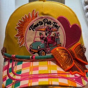 Op de afbeelding: Een gele baseballpet met een ronde patch van een flamingo in een golfkarretje en de tekst Time to Par-Tee. De pet is voorzien van een oranje zon-patch, een roze hart, een turquoise kralenrand en een grote oranje strik met lovertjes op de klep.