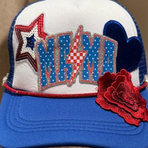 May include: A blue and white trucker hat with the word "MIMI" in a sparkly, patriotic design. The hat features a red star, a blue heart, and a red flower. The brim is blue, and the mesh is white.