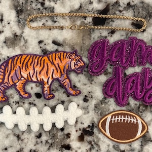 May include: A gold-tone chain, an embroidered tiger patch in orange and purple, a purple glitter script patch reading game day, a white felt football lace patch, and a brown felt football patch with white stitching details.