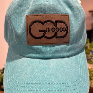 May include: A turquoise blue baseball cap with a brown leather patch that says "God is Good" in black lettering.