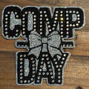 May include: A decorative patch featuring the text COMP DAY in black letters with silver glitter borders and accents. A large silver glitter bow is positioned between the two words. The entire design is outlined with clear rhinestones.