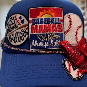 Op de afbeelding: Een blauwe baseballpet met meerdere patches. Eén patch leest "Busy Raising Ballers", een andere zegt "Baseball Mamas Always Salt", en een derde is een honkbal. Een rode paillettenstrik is bevestigd.