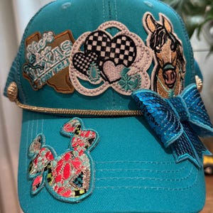 May include: A turquoise baseball cap featuring a gold rope trim, a blue sequined bow, and embroidered patches including a brown Texas shape with text, a black and white checkered heart, a horse head, and a colorful patterned cactus shape.