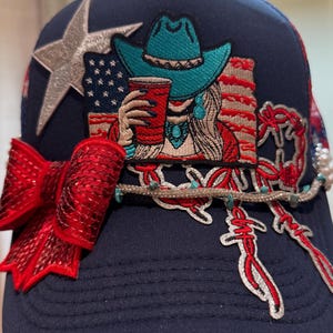May include: Navy blue baseball cap with a red sequined bow and a silver star. The cap features an embroidered design of a woman in a turquoise cowboy hat holding a red cup, with an American flag background. A decorative beaded wire with charms is also present.