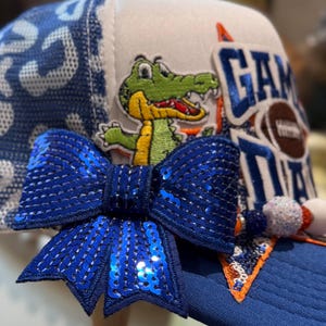 May include: A white and blue trucker hat featuring a large blue sequined bow, an embroidered green alligator patch, and a patch with the text GAME DAY and a football graphic. The side panel has a blue and white leopard print mesh pattern.
