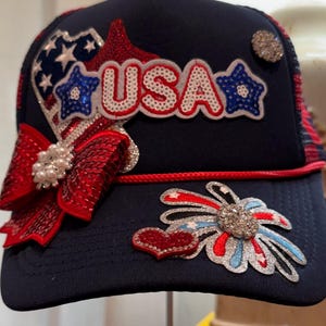 Puede incluir: Gorra de camionero azul marino con adornos patrióticos. Presenta la palabra "USA" en letras de lentejuelas, un lazo rojo de lentejuelas y detalles de estrellas y corazones. La gorra tiene una parte trasera de malla a rayas rojas y blancas.