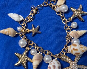 May include: Gold-toned charm bracelet featuring seashells, starfish, and faux pearls. The bracelet has a delicate chain design with various seashell shapes, including spiral and conch shells, and small, round pearls. The starfish charms add a touch of nautical flair.