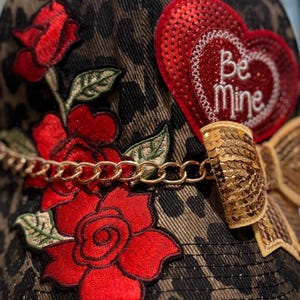 May include: A baseball cap with a leopard print pattern, adorned with red embroidered roses, a gold chain, and a sequined heart that says "Be Mine". A gold sequined bow adds a decorative touch to the cap.