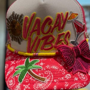 Op de afbeelding: Een witte en rode truckerpet met de woorden "VACAY VIBES" in roze en geel. De pet heeft een paillettenstrik, een palmboom en een cocktailafbeelding. De rand heeft een rood paisley patroon.