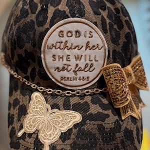 May include: A baseball cap with a brown and black leopard print pattern. The cap features a gold butterfly and a gold sequined bow. A circular patch with the text "GOD IS within her SHE WILL not fall" is also on the cap.