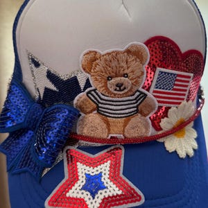 May include: A blue and white trucker hat with patriotic embellishments. Features a brown teddy bear wearing a striped shirt, a sequined bow, and a red, white, and blue star. Includes a heart with an American flag.