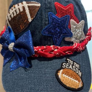 May include: Denim baseball cap decorated with a sequined football, blue bow, red and silver stars, and a red bandana. The cap also features a patch that says "Tis the Season" and a football.