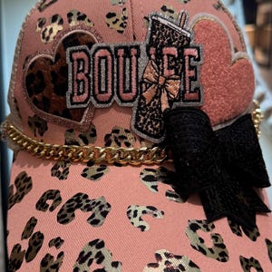 Może przedstawiać: Różowa czapka bejsbolówka z wzorem w panterkę i złotym łańcuszkiem. Czapka ma napis "BOUJEE" w srebrnych literach, czarną kokardę oraz naszywki w kształcie serca i napoju.