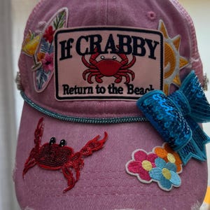 Puede incluir: Gorra de béisbol rosa con parte trasera de malla y visera curva. La gorra presenta parches bordados de un cangrejo, flores, un cóctel y un sol. El texto "If Crabby Return to the Beach" se muestra en un parche.