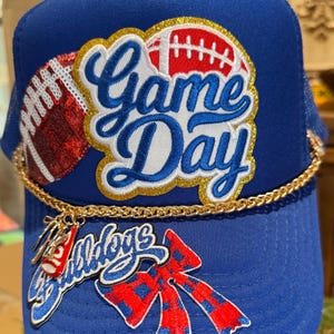 May include: A blue trucker hat with a gold chain and charms. The hat features the words "Game Day" in white and gold, along with football graphics. A charm reads "Bulldogs" with a red and blue bow.