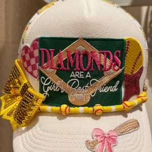 Puede incluir: Gorra de béisbol blanca con parte trasera de malla, con un parche verde con el texto "Diamonds Are a Girl's Best Friend". Adornada con un lazo de lentejuelas amarillo, abalorios con temática de béisbol y un lazo rosa con un bate.