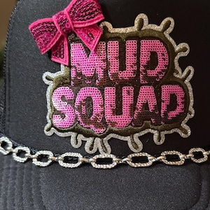 Puede incluir: Gorra de camionero negra con un lazo de lentejuelas rosa y un parche de lentejuelas "MUD SQUAD". El parche tiene un contorno marrón con detalles de purpurina plateada. Una cadena plateada está unida a la visera.