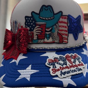May include: A white and blue trucker hat featuring a front patch of a person in a turquoise cowboy hat holding a red cup against an American flag. The hat includes a red sequin bow, a blue sequin star, a silver chain, and a patch reading She loves Jesus and America too.