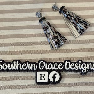 May include: A pair of dangle earrings with silver-colored studs and black and white leopard print tassels. The earrings are displayed on a striped background. The image also includes the text "Southern Grace Designs".