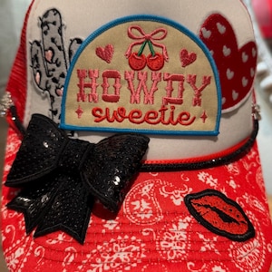 May include: A trucker hat with a white front and red mesh back. It has a black sequined bow, a cactus patch, and a patch with cherries and the words "Howdy Sweetie". The red brim has a paisley pattern.