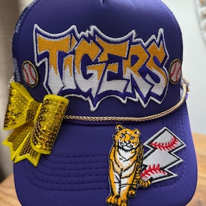 May include: Purple baseball cap with the word "TIGERS" in yellow and white lettering. The cap features a yellow sequined bow, baseball patches, and a tiger patch. A gold rope trim accents the cap.